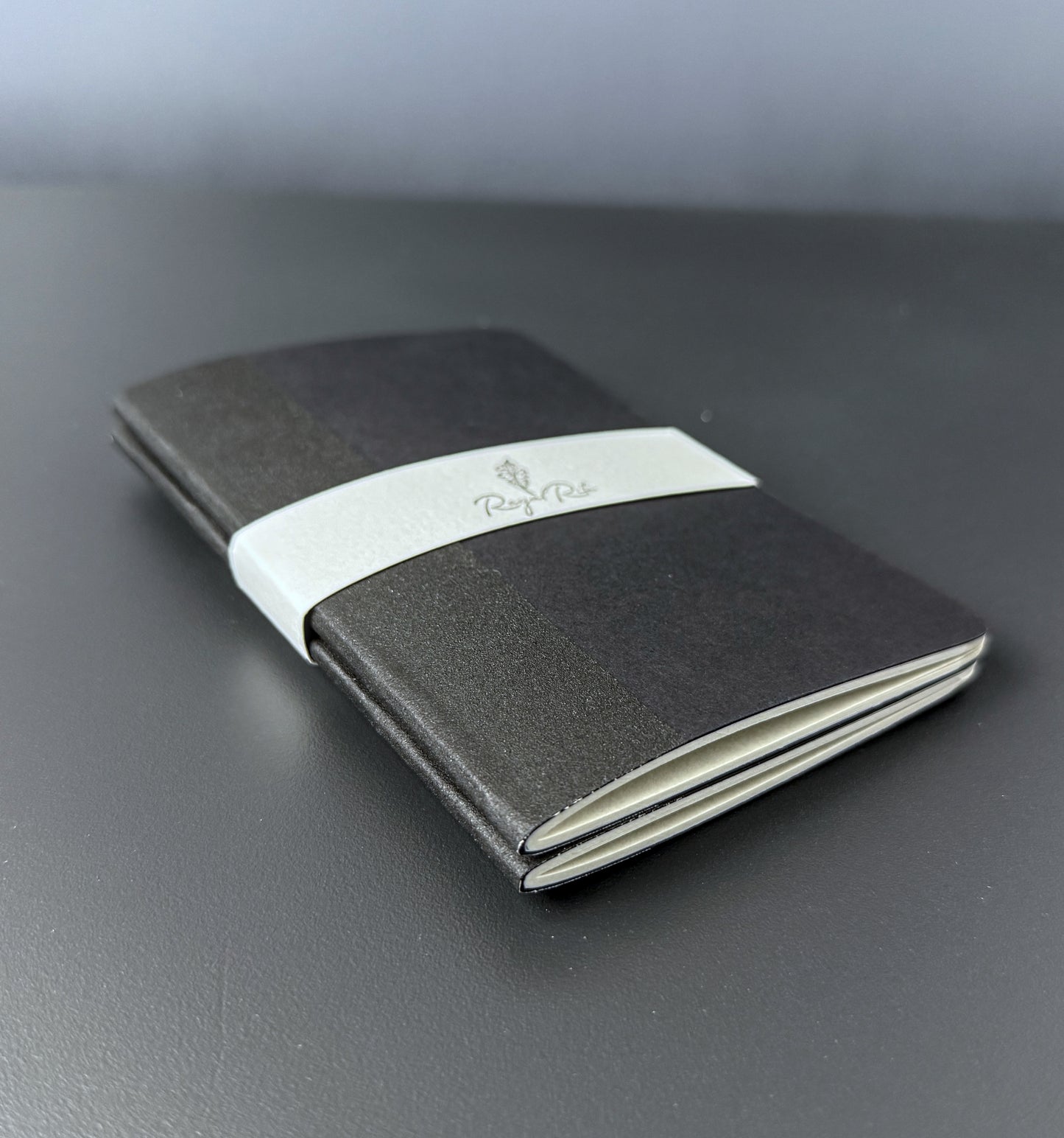 A black pocket notebook cover with a white paper band around the middle, featuring the 'Royal Rite' logo, resting on a dark gray surface.
