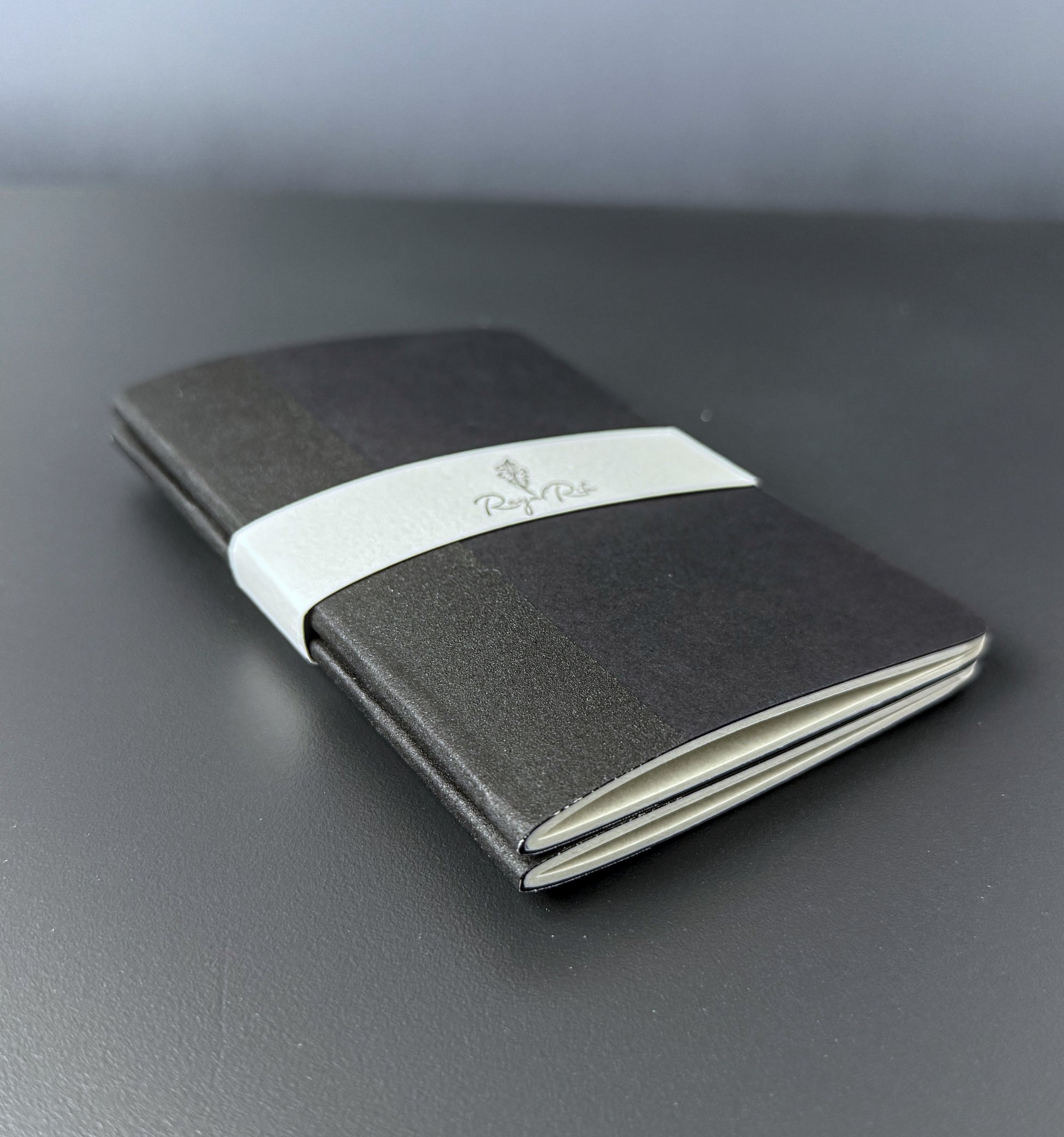 A black pocket notebook cover with a white paper band around the middle, featuring the 'Royal Rite' logo, resting on a dark gray surface.