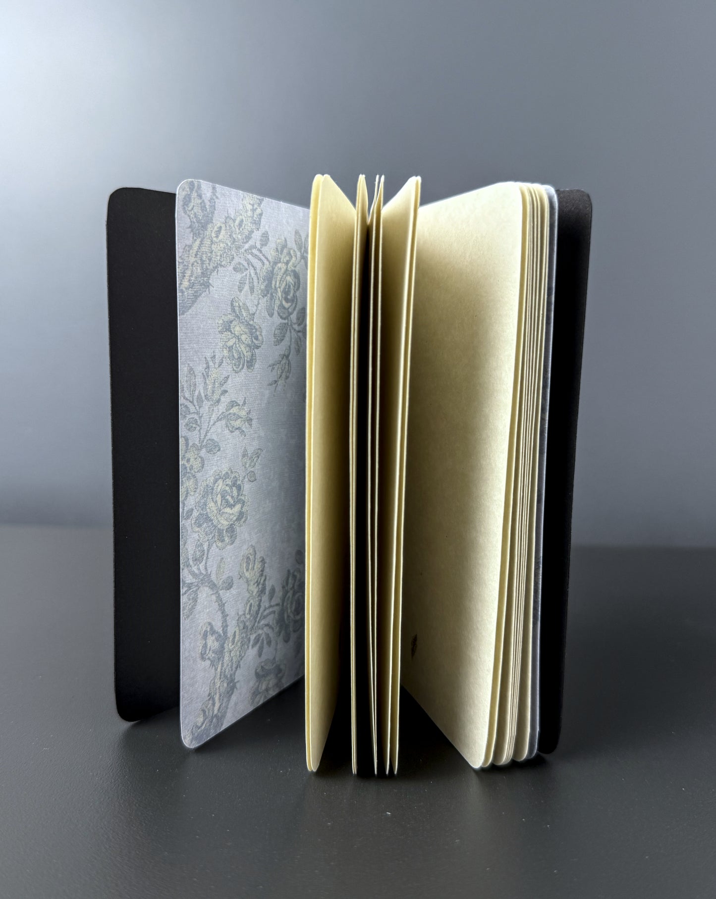 An open notebook or journal standing upright on a dark surface against a gray background. The notebook has a black cover, cream-colored pages, and a floral-patterned paper insert on the left side.