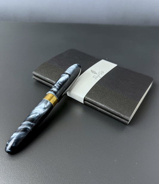A black and white marbled fountain pen with a gold band, placed next to a dark gray notebook with a white paper band around it, on a dark gray surface.